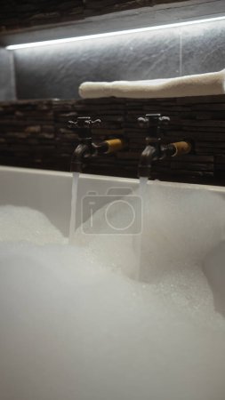 Une baignoire luxueuse remplie de mousse sous des robinets vintage et un éclairage atmosphérique.