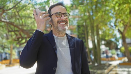 Photo for Smiling bearded man listening to a voice message on a smartphone in a sunny park - Royalty Free Image