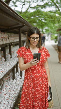 Cheerful beautiful hispanic woman in glasses happily using smartphone at tokyo's lucky gotokuji temple, connecting online with a radiant smile