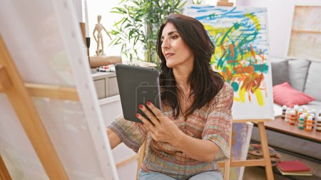 Téléchargez les photos : Une artiste hispanique d'âge moyen tenant une tablette dans un studio bien éclairé avec des peintures colorées et des fournitures d'art autour d'elle. - en image libre de droit