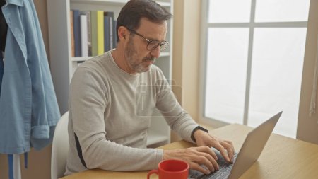 A mature man with a beard working on a laptop in a bright home office space.