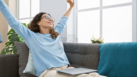 Photo for Happy middle-aged woman stretching in living room with laptop, plants, glasses, and homey decor. - Royalty Free Image