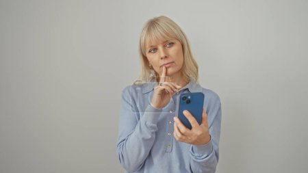 Foto de Mujer rubia pensativa sosteniendo smartphone sobre fondo blanco, expresando contemplación y curiosidad. - Imagen libre de derechos