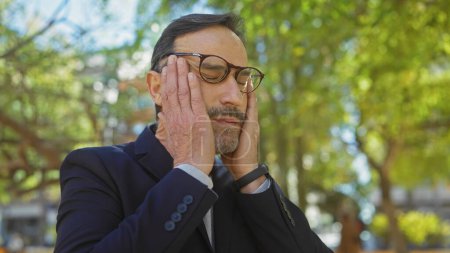 A stressed mature man in glasses holds his face in a park, embodying frustration or headache in an outdoor setting.