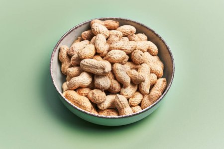 A bowl full of unshelled peanuts on a textured green background, portraying a simple yet captivating still life composition.
