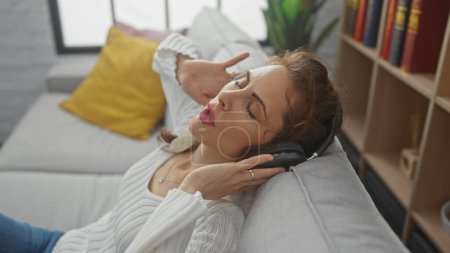 Foto de Una joven relajada disfrutando de la música con auriculares mientras está tumbada en un sofá en una acogedora sala de estar. - Imagen libre de derechos