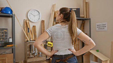 Photo for Confident young woman with ponytail stands in carpentry workshop, hands on hips, tools and wood background. - Royalty Free Image
