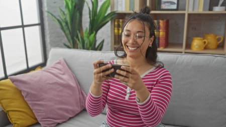 Foto de Una mujer hispana alegre se relaja en casa, casualmente absorta en su teléfono inteligente en un acogedor sofá adornado con cojines de colores. - Imagen libre de derechos