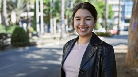 Téléchargez les photos : Jeune femme hispanique souriante en veste de cuir debout à l'extérieur dans un parc urbain ensoleillé. - en image libre de droit
