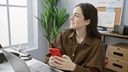 Une jeune femme caucasienne en chemisier marron sourit en utilisant un smartphone dans son espace de travail organisé.