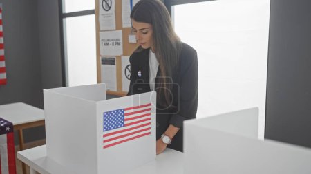 Mujer hispana joven en un traje que vota atentamente en un centro electoral americano con bandera