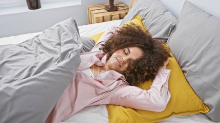 Téléchargez les photos : Une jeune femme hispanique aux cheveux bouclés dort paisiblement dans une chambre confortable, mettant en valeur la détente à la maison. - en image libre de droit