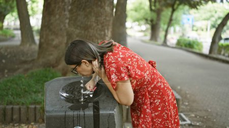 Preciosa mujer hispana saciando la sed con refrescante bebida de la fuente del parque, sus gafas brillando con el chapoteo del verano! hermoso retrato al aire libre afirmando opciones saludables e hidratantes.