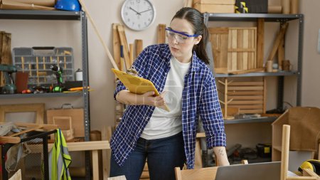 Femme caucasienne avec des lunettes de protection examine les plans dans un studio de travail du bois entouré d'outils