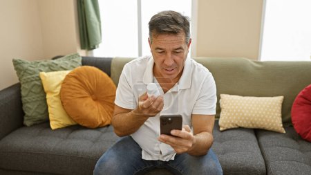 Middle-aged man examining pill bottle while using smartphone in cozy living room.