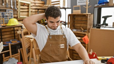 Foto de Un hombre pensativo con barba en un taller de carpintería, rodeado de herramientas y muebles de madera. - Imagen libre de derechos