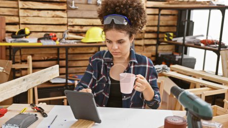 A young hispanic woman with curly hair sips a mug of coffee while studying a digital tablet in a carpentry workshop.