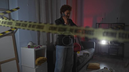 Foto de Una mujer afroamericana preocupada en una llamada telefónica en una sala de escena del crimen débilmente iluminada acordonada por cinta de la policía - Imagen libre de derechos