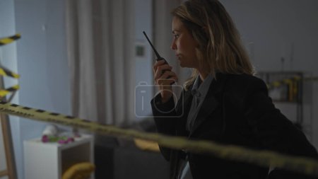 Photo for A focused blonde woman wearing a black suit communicates on a handheld radio at an indoor crime scene cordoned with yellow tape - Royalty Free Image