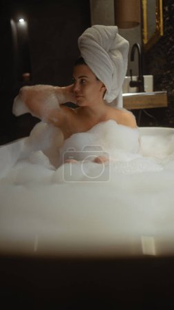 Téléchargez les photos : Une jeune femme profite d'une baignoire en mousse luxueuse dans un cadre de salle de bain à la maison bien aménagé. - en image libre de droit