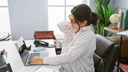 Foto de Una mujer hispana profesional experimenta dolor de cuello mientras trabaja en una computadora portátil en un entorno de oficina moderno - Imagen libre de derechos