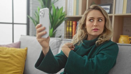 Téléchargez les photos : Une jeune femme caucasienne prend un selfie à l'intérieur avec son smartphone, mettant en valeur une configuration confortable de salon. - en image libre de droit