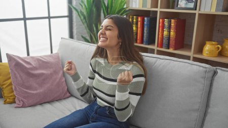 Téléchargez les photos : Jeune femme hispanique excitée célébrant à la maison sur un canapé confortable. - en image libre de droit