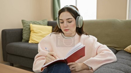 Téléchargez les photos : Une jeune femme concentrée étudie dans son confortable salon, écouteurs allumés, notes dans un carnet rouge. - en image libre de droit