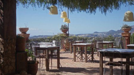 Terraza exterior en un restaurante mediterráneo con vistas panorámicas, mesas y lámparas colgantes.