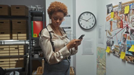 Téléchargez les photos : Femme afro-américaine détective avec téléphone dans une salle de police, tableau des preuves en arrière-plan - en image libre de droit