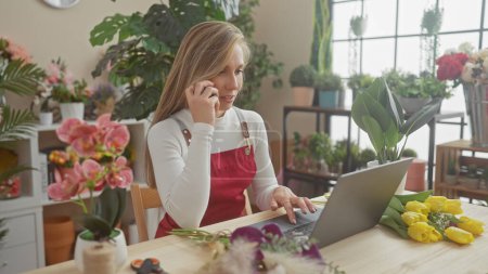 A blonde woman florist in a red apron using a laptop and phone amidst vibrant flowers inside a florist shop.