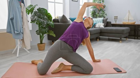 Photo for Blonde woman stretching on a yoga mat in a modern living room, embodying wellness and an active lifestyle. - Royalty Free Image