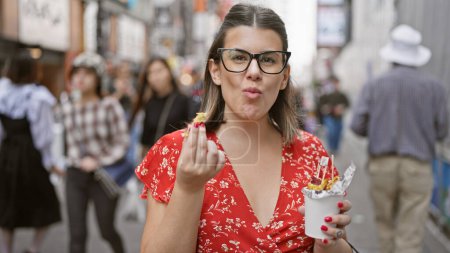 Stunning hispanic woman savors sweet potato snack in her eyeglasses on the buzzing osaka's dotonbori street