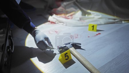 Foto de Un investigador forense examina un martillo ensangrentado en una escena del crimen con marcadores de evidencia en una habitación con poca luz. - Imagen libre de derechos