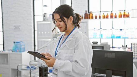 Foto de Mujer hispana joven con bata blanca usando tableta en laboratorio moderno, enfocada y profesional. - Imagen libre de derechos