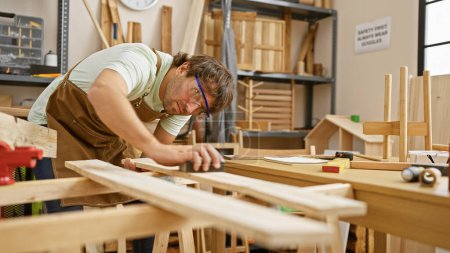 Photo for A young bearded man sands wood in an indoor carpentry workshop, wearing safety glasses and a brown apron. - Royalty Free Image