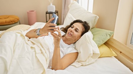Eine reife hispanische Frau entspannt sich in einem Schlafzimmer mit einem Smartphone, während sie bequem im Bett liegt, eingetaucht in eine morgendliche Routine.
