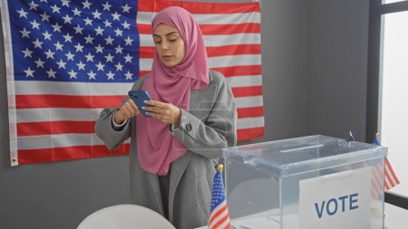 A young woman in a hijab using a smartphone by an american flag and a voting box indicates electoral engagement.