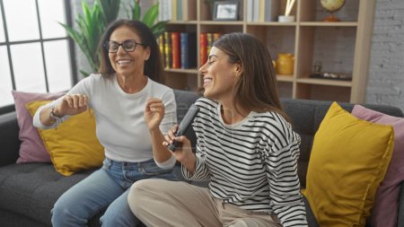 Foto de Madre e hija riendo juntas, sosteniendo un mando a distancia como un micrófono en una acogedora sala de estar - Imagen libre de derechos