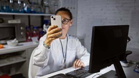 Foto de Una científica madura en un laboratorio examina un teléfono inteligente mientras trabaja en una computadora, rodeada de equipos de laboratorio. - Imagen libre de derechos