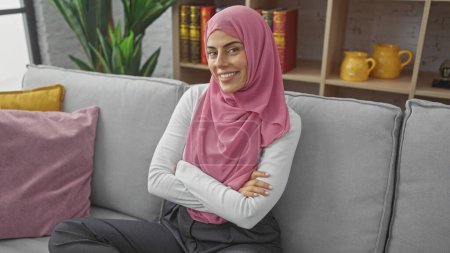 Mujer joven sonriente con los brazos cruzados usando hijab sentado en un sofá gris