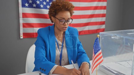 Afroamerikanerin im blauen Blazer macht sich drinnen Notizen, im Hintergrund eine amerikanische Flagge, die eine professionelle Wahlatmosphäre darstellt.