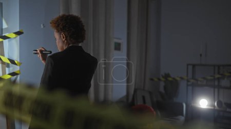 Téléchargez les photos : Une femme afro-américaine professionnelle enquête sur une scène de crime dans une pièce intérieure faiblement éclairée avec un ruban adhésif - en image libre de droit