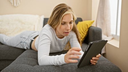 A young blonde woman in casual attire lies on a couch, absorbed in using her digital tablet in a modern living room.
