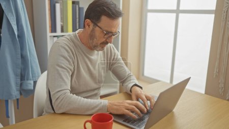Mature bearded man typing on laptop at home office with glasses and red mug.