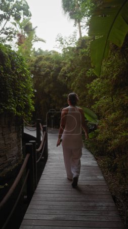 Foto de Una mujer camina por un sendero de madera rodeado de exuberante vegetación tropical, personificando el ambiente sereno del resort en bali. - Imagen libre de derechos