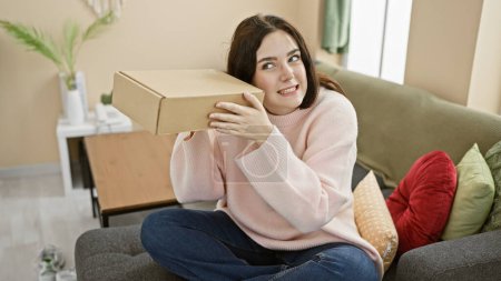 Curious young woman listens to a mystery cardboard box while sitting comfortably in her modern living room.