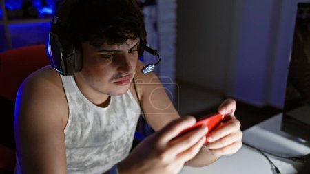 A young man plays a handheld game in a dimly lit gaming room at night, showcasing concentration and entertainment.
