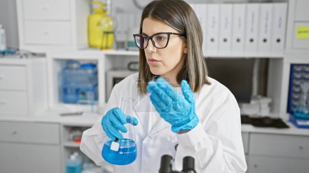 Foto de Científica hispana examina líquido azul en frasco en laboratorio de interior - Imagen libre de derechos
