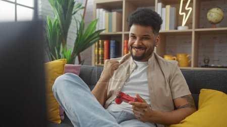 Téléchargez les photos : Beau homme noir appréciant les jeux vidéo à la maison, visiblement content et détendu dans un environnement confortable salon. - en image libre de droit